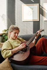 Young woman playing guitar while resting on the sofa during her leisure time at home