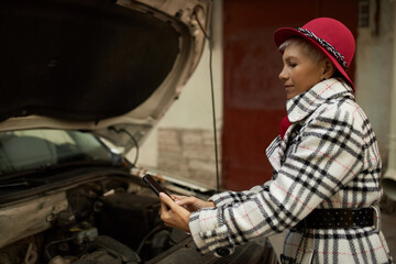 Outdoor image of business woman in top coat and hat taking picture of broken car engine under...