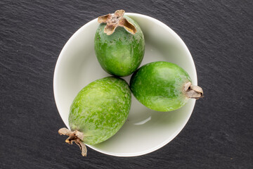 Three fragrant sweet feijoa fruits with a white saucer on a slate stone, close-up, top view.