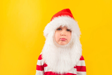 Unhappy child toddler in a Santa hat with red gift , yellow background. Holidays, Traditions, Christmas concept.
