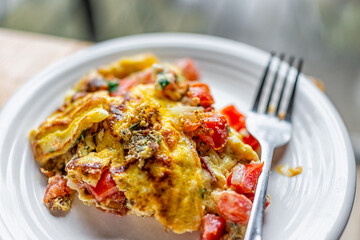 Macro closeup of homemade white plate with cooked ready to eat omelette with tomatoes, cilantro and red peppers vegetables on wooden kitchen table with fork