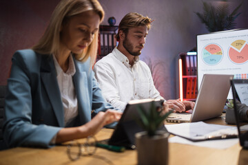 Caucasian man and woman in formal wear working on modern gadgets while sitting at desk. Two colleagues cooperating for common project during evening time.