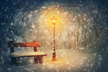 Wintertime night scene in the park. Shining street lamp near a wooden bench covered with snow. Wonderful winter holidays seasonal background. Christmas Eve and New year magic Noel atmosphere - Powered by Adobe