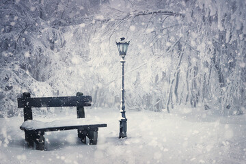 Winter time scene with a wooden bench and street lamp covered with snow under a snowfall in the...