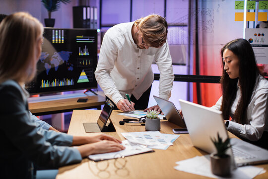 Group of competent traders sitting together at office desk with various graphs and charts and working on gadgets. Multiracial partners during working process indoors.