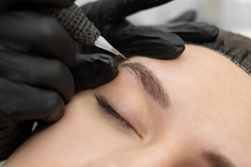 Concept of microblading eyebrows. The cosmetologist performs the procedure of permanent makeup of the eyebrows in close-up. - obrazy, fototapety, plakaty