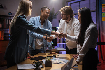 Team of multicultural software testers shaking hands while standing near table with various gadgets and papers. Meeting with a customer. Successful cooperation.