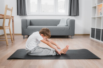 Boy child makes a forward bend from a sitting position on the floor with straight legs, wearing a white t-shirt and gray pants.