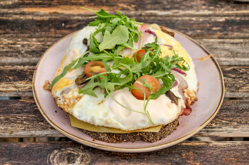 Plate with fried eggs, bacon, cheese and champignons on brown bread, garnished with tomato and rucola, on a rough wooden table