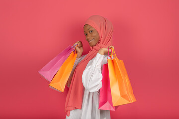 Concept of seasonal sales and shopaholic. Smiling Muslim woman in profile holding many bright shopping bags looking at the camera on a pink background.