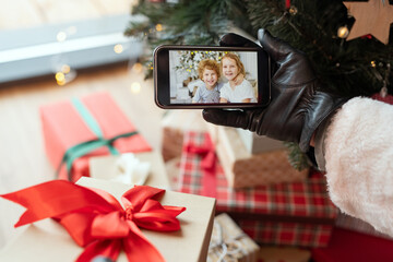 Naklejka na ściany i meble Hand in black leather glove holding smartphone with photo of kids on screen against Christmas presents and tree
