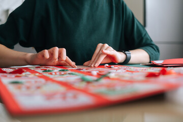 Close-up of unrecognizable young woman gluing envelopes on board with gifts for children making Christmas advent calendar at home, selective focus, blurred background. Preparing for xmas, new year. - obrazy, fototapety, plakaty