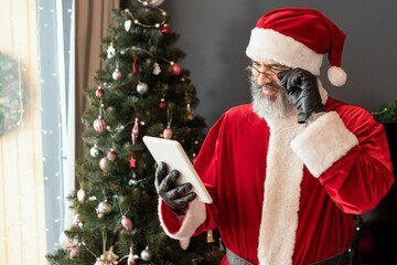 Content Santa Claus in black gloves adjusting eyeglasses while reading internet news on tablet in living room with Christmas tree
