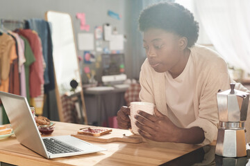 African young woman having breakfast with toast and jam and watching something on laptop while sitting at the table in her room