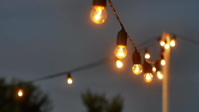 Festival decorative string lights hang and glow outdoors at night. Warm lighting, vintage garland of lamps or glass lantern, electric bulbs shining, decoration for holiday.