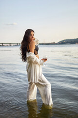 Sexy young brunette in a black bikini and white suit with a glass of wine posing on the beach. High quality photo