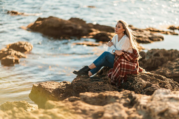 Attractive blonde Caucasian woman enjoying time on the beach at sunset, sitting in a blanket and looking to the side, with the sunset sky and sea in the background. Beach vacation.