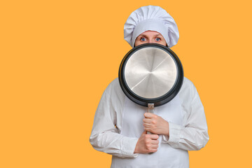 Frightened woman chef closes with a frying pan on yellow studio background, copy space