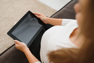 Joyful pregnant woman having fun with a tablet while sitting on sofa at home.