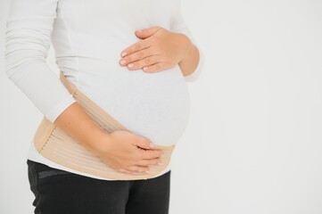 Close up of pregnant woman putting on a bandage at gray background with copy space. Orthopedic abdominal support belt concept.