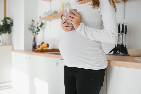 Picture of beautiful casually dressed young brunette female expecting baby, having morning tea in modern stylish kitchen, smiling. Anticipation, expectation, childbearing and motherhood concept