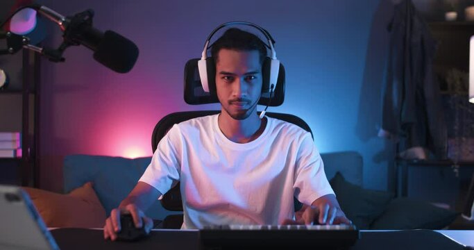 Young Asian man turn chair around, start playing online computer video game, colorful lighting broadcast streaming live at home. Gamer lifestyle, E-Sport online gaming technology concept