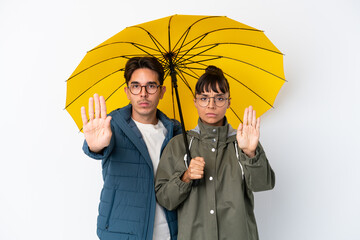 Young mixed race couple holding an umbrella isolated on white background making stop gesture denying a situation that thinks wrong