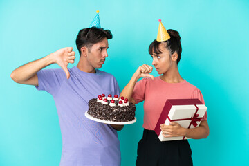 Young mixed race couple holding birthday cake and present isolated on blue background showing thumb...