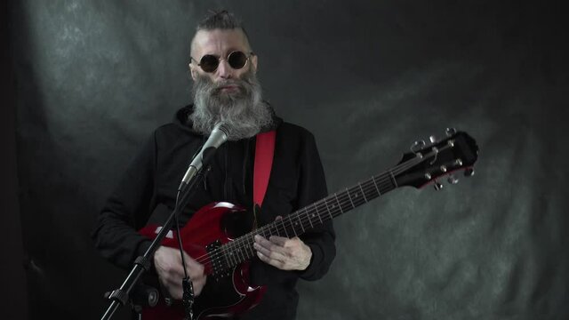 Bearded hairy rockstar in retro round sunglasses and black hoodie with shaved temples plays on red electric guitar on stage with black background