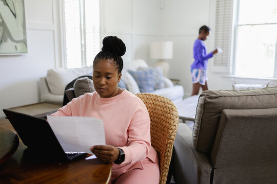 Woman using laptop and reading document at table in living room, teenage daughter in background