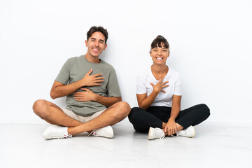 Fototapeta na wymiar Young mixed race couple sitting on the floor isolated on white background smiling a lot while putting hands on chest