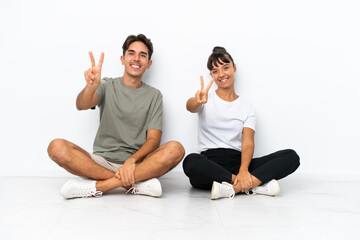 Fototapeta na wymiar Young mixed race couple sitting on the floor isolated on white background smiling and showing victory sign