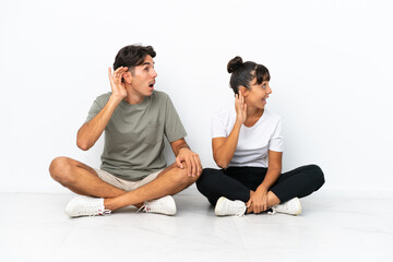 Fototapeta na wymiar Young mixed race couple sitting on the floor isolated on white background listening to something by putting hand on the ear