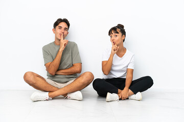 Young mixed race couple sitting on the floor isolated on white background showing a sign of closing mouth and silence gesture