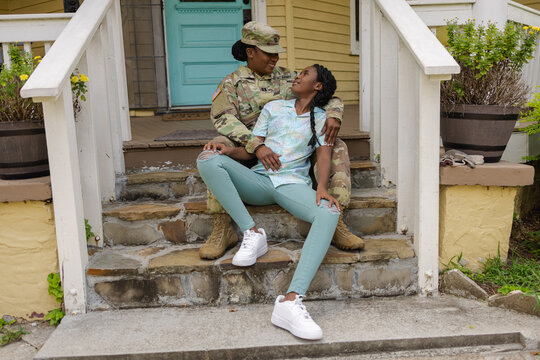 Happy military woman in camouflage uniform sitting with teenage daughter on steps in front of house