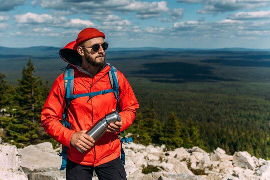 A bearded traveler with a backpack on the top of a mountain. Portrait of a traveler in a red cap and sunglasses. A tourist with a backpack stands against the background of a mountains. Copy space