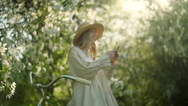 Portrait of Blonde Woman in Hat Enjoy Takes Pictures at on Smartphone Device of White Apple Blossoms. Garden With Blooming Trees. The Concept of Beauty, Health, Freshness and Naturalness. Close up