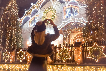 Rear view of female who making heart shape in front of decorated house whit Christmas lights. Holidays concept