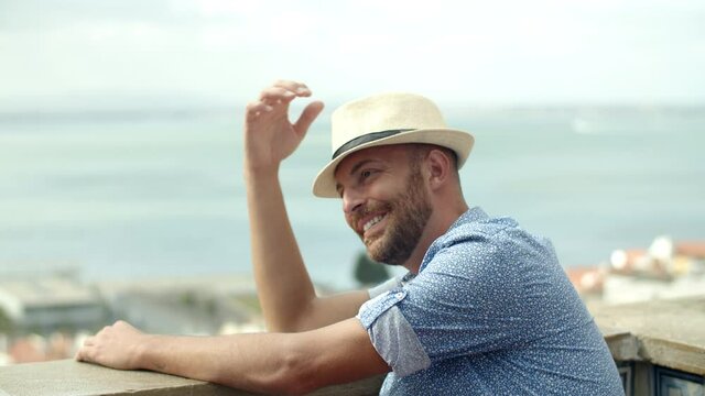 Slow motion of happy man sitting on observation tower. Side view of laughing male person wearing hat admiring view of city from above, enjoying summertime in Europe. Emotion, landscape concept