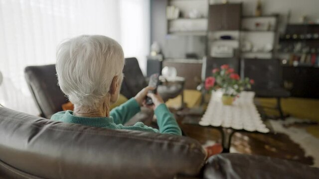 Medium shot - Slow motion of the backside of a grandmother's head, while she is sitting in a living room with her legs crossed and she is changing the TV programs, with red roses on the table and brow