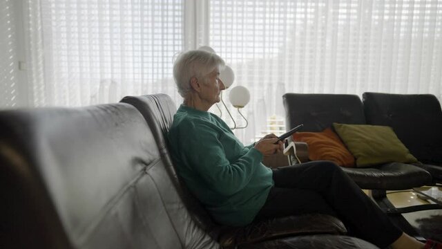 Slow motion - An old woman sitting in a living room and changing TV programs with a remote