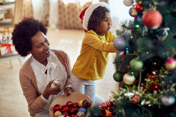 Happy African American mother and daughter decorate Christmas tree in their living room.