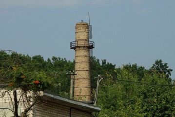 Fototapeta premium one old tall brown brick water tower on the street against a background of sky and green trees