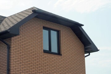 brown brick attic of a private house with one window under a black tiled roof against a blue sky