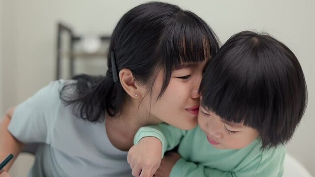 Mom kissing son and teaching how to painting with crayon color on book or doing homework. Happy asian boy painting with crayon with his mother in living room at home. Family having fun concept.