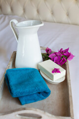 A white jug of water, a towel and soap are on a wood tray on a white bed. The theme is morning hygiene procedures. Retro style. Vertical image.
