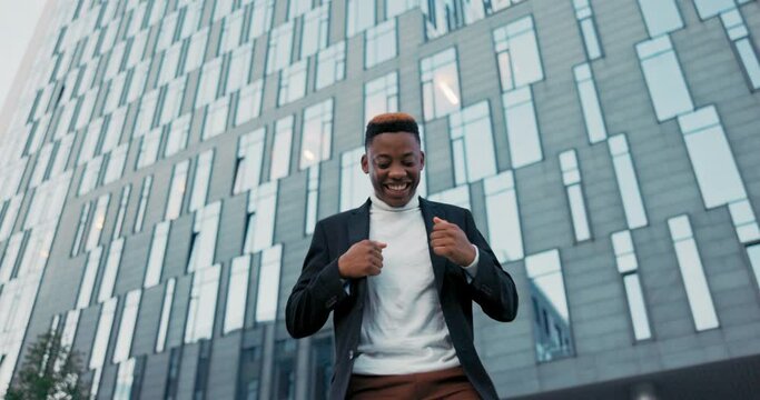 Joyful, positive man dressed in suit stands outside in front of glass building of company, corporation, office worker received good news happy dancing waving hands satisfaction promotion, salary