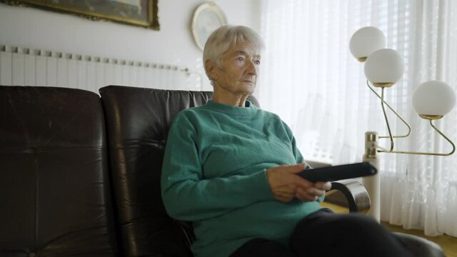 Slow motion - Zooming in on a grandmother, holding a TV remote and watching television, while sitting in a living room on a leather couch, with a big window with blinds in the background, and some pic