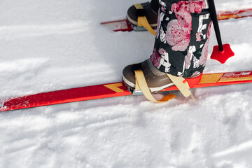 Close-up of ski mounts. Red skis attached with ribbons to winter children shoes, child on winter walk, snowy background. 
