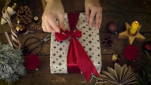 Merry Christmas! Hands opening stylish christmas gift with red ribbon on rustic wooden table with festive holiday decorations. Top view. Happy Holidays! Atmospheric Xmas footage